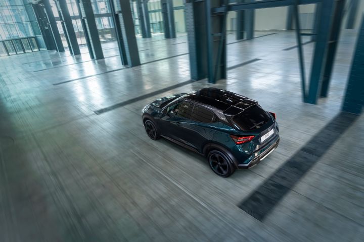 Top rear three-quarter view of the Nissan JUKE Pulse driving through a large indoor industrial space, highlighting its contrasting black roof and compact crossover design