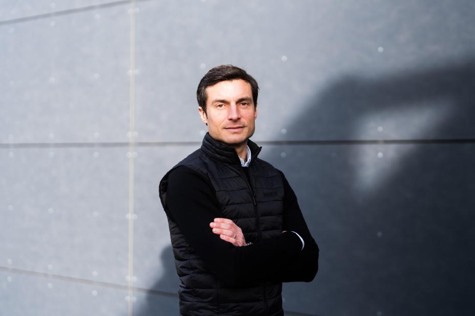 Portrait of Bruno Spengler, with his arms crossed, posing in front of a grey brick wall.