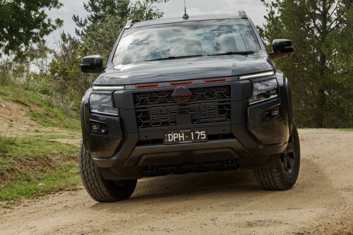 2026 Nissan Navara PRO-4X finished in Boulder Grey frontal on a rutted track, red‑highlighted grille and lower protection visible beside pine trees.