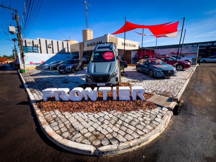 Imagem externa de um estande da Nissan com diversos carros expostos. Em destaque, uma picape Nissan Frontier cinza posicionada sobre uma área central com pedras, com letras tridimensionais brancas formando a palavra “FRONTIER”. Ao fundo, há mais veículos, o estande com o logotipo da Nissan e uma estrutura com lonas vermelha e branca que fornecem sombra parcial.