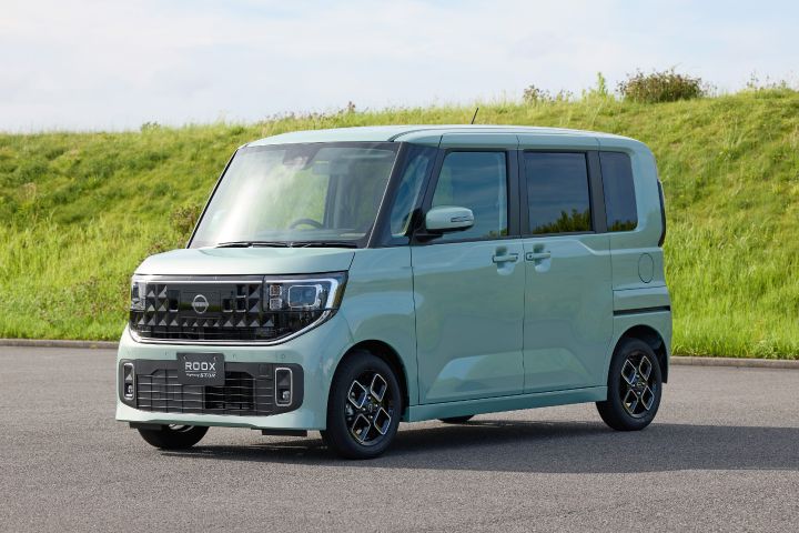 The new Nissan Roox kei car in celadon green is parked against a backdrop of green grass. Its boxy shape stands out while blending harmoniously with the natural surroundings, creating a calm and balanced impression.