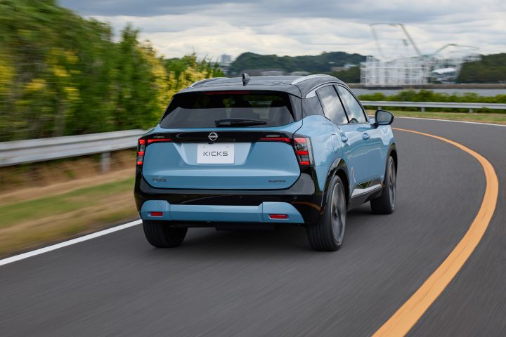 青みがかったキックスがカーブした道路を右方向に走行する後方視点のカット。車両は後ろ姿で右向きに進み、ガードレールや緑の植生が沿道に見える。