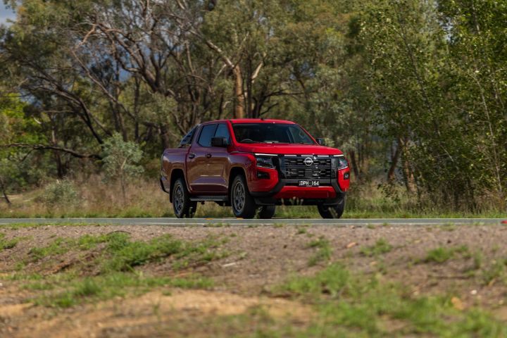 Front three‑quarter motion shot of a 2026 Nissan Navara ST‑X Outback Red dual cab driving on a sealed rural road bordered by tall trees.