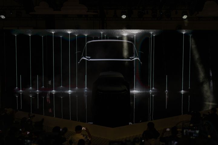 In a dark stage setting, the all-new Elgrand is positioned at the center, covered with a black cloth, while illuminated contour lines of the vehicle appear in the background. In the foreground, members of the press are gathered, taking photos with cameras.