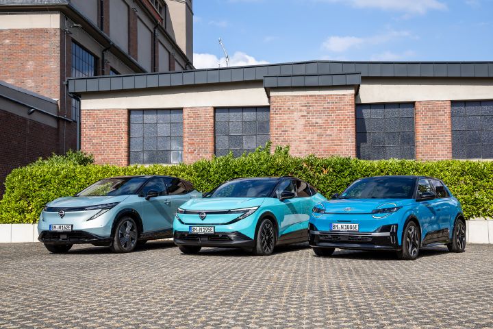 Vor einem Backsteingebäude und einer grünen Hecke stehen nebeneinander die drei Nissan EV-Modelle Nissan Ariya, Nissan Leaf und Nissan Micra, Ansicht von schräg vorne und leicht unten