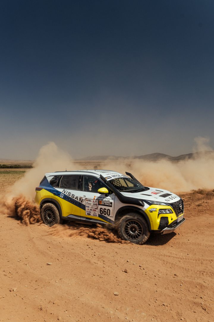 Fahraufnahme des Nissan X‑Trail Rennwagens während der Baja Aragon Rallye 2024, das Auto wirbelt Sand auf und der Beifahrer zeigt ein Peace-Zeichen aus dem Fenster