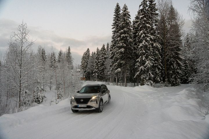 Silberner Nissan X‑Trail e‑Power e‑4ORcE fährt auf schneebedeckter Straße mit verschneiten Bäumen auf beiden Seiten, 3/4 Front