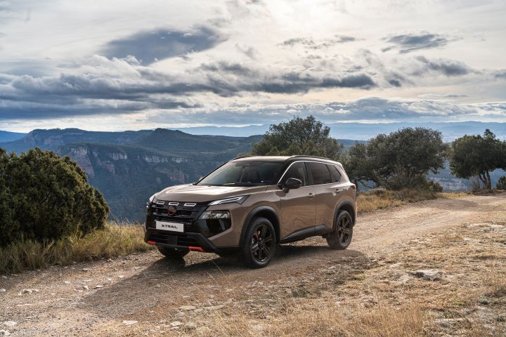 Der neue 2026 Nissan X‑Trail N-Trek in der Karosseriefarbe „Baja Storm“ steht auf einer unbefestigten Straße in einer bergigen Landschaft bei bewölktem Himmel. Das Fahrzeug verfügt über schwarze Akzente, magmarote Zierdetails und robuste Reifen, die seine Geländegängigkeit und sein abenteuerlustiges Design unterstreichen.