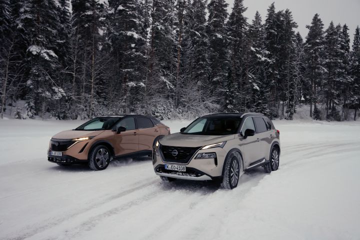 Elektroauto Nissan Ariya e‑4ORCE in Akatsuki Copper mit Dach und Außenspiegel in Pearl Black und silberner Nissan X‑Trail e‑Power e‑4ORCE fahren nebeneinander auf schneebedeckter Straße mit verschneitem Wald im Hintergrund, 3/4 Front