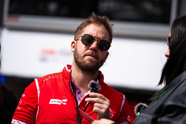Sam Bird trägt sein rotes Team-Polohemd und seine Weste sowie seine Sonnenbrille und hält ein kleines Mikrofon in der linken Hand.
