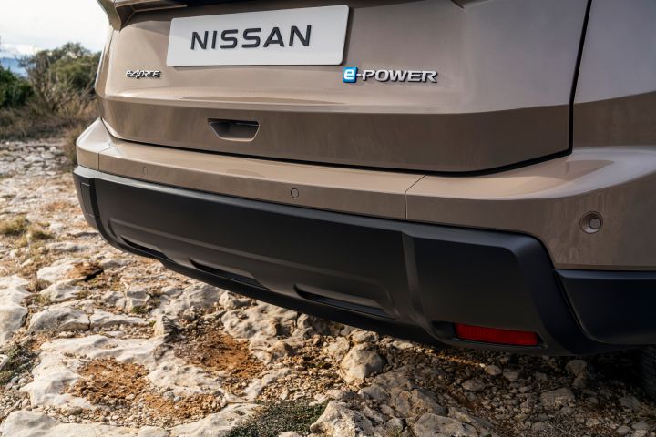 Der neue 2026 Nissan X‑Trail zeigt eine Nahaufnahme der hinteren Stoßstange und der unteren Heckklappe eines Nissan-Fahrzeugs mit „e‑POWER”-Emblem, das auf felsigem Gelände geparkt ist. Das Bild hebt Designdetails wie die Karosseriefarbe „Baja Storm”, die schwarze untere Stoßstange und den magmaroten Reflektor auf der rechten Seite hervor.