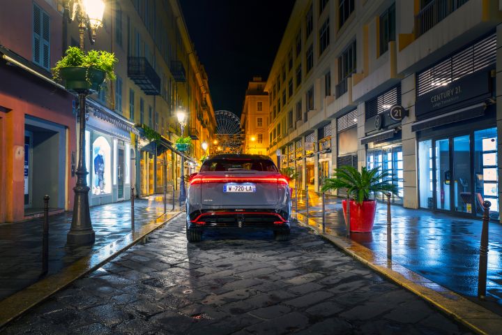 Vue arrière d'une Nissan Ariya NISMO bleu-gris avec des accents de design rouge, qui roule de nuit dans une rue commerçante étroite et éclairée