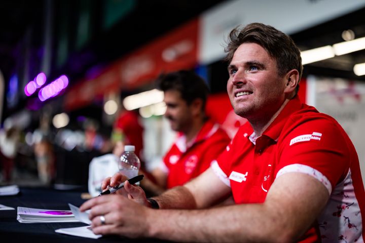 Oliver Rowland, der ein rotes Team-Trikot trägt, sitzt an einem Tisch, hält einen Stift in der Hand und blickt lächelnd nach vorne. Im Hintergrund ist Norman Nato in ähnlicher Kleidung und bei unscharfem Licht zu sehen.