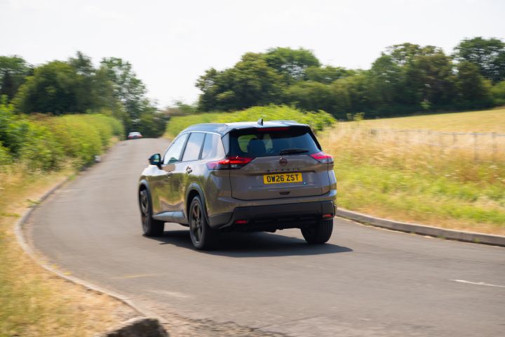 Nissan X‑Trail driving along a country road, shown from the rear, with a field to the right .