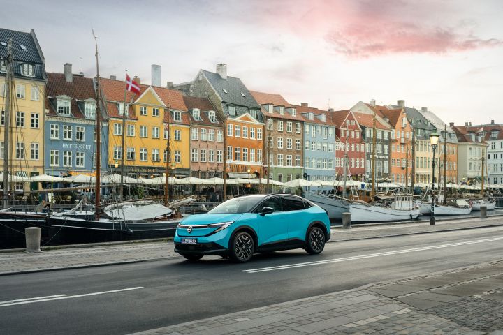 Hinter einem Kanal mit Segelbooten stehen mehrere farbenfrohe Stadthäuser in Kopenhagen, im Vordergrund fährt ein türkiser Nissan LEAF