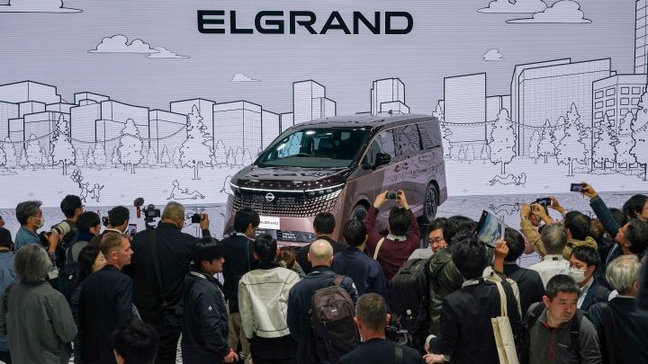 On the stage, the all-new Elgrand in Fuji Dawn and Shigoku two-tone colors is positioned diagonally, facing slightly to the left. The background features a white-themed screen displaying an urban line drawing and the word ELGRAND. In the foreground, members of the press are gathered, taking photos with their cameras.”