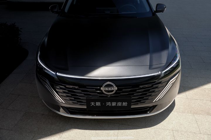 A dark gray new Teana sedan parked on a paved surface, facing directly forward. The design highlights a wide V-motion style grille and slim LED headlights. The background is minimalistic, with soft shadows cast on the ground and no visible buildings or urban elements. The overall tone is subdued, emphasizing the car’s front-end styling.