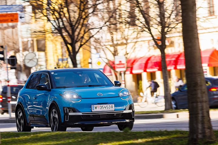 Blick von schräg vorne auf einen leuchtend blauen Nissan Micra mit schwarzem Kontrastdach, der eine belebte Straße entlangfährt, im Hintergrund große rote Sonnenmarkisen vor einem hellen Gebäude