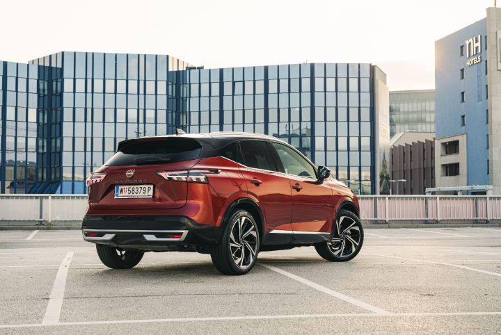Ein Nissan Qashqai in Fuji Sunset Red steht auf einem Parkplatz vor einem verglasten Hotel-Hochhaus, 3/4 Heck