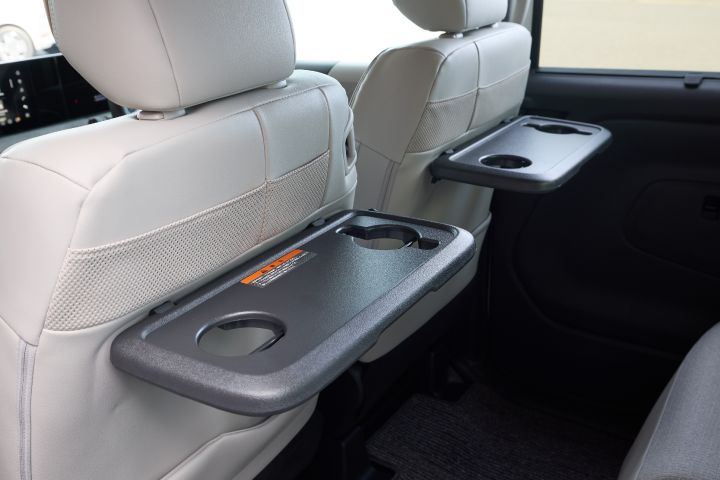 The foldable tray tables attached to the backrests of the rear seats in the new Nissan Roox kei car. Each tray includes two cup holders and can be folded away when not in use. The background shows part of the car interior.