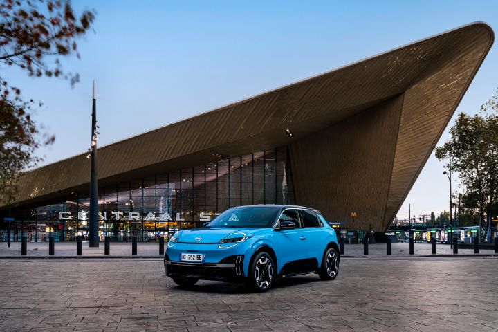 3/4-Frontansicht eines blauen Nissan Micra, der vor der metallisch reflektierenden, spitz zulaufenden Fassade des Hauptbahnhofs von Rotterdam steht