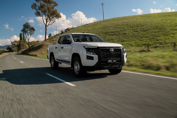 2026 Nissan Navara SL dual cab ute finished in Alpine White driving from a front three-quarter angle beside grassy hills and roadside fencing.