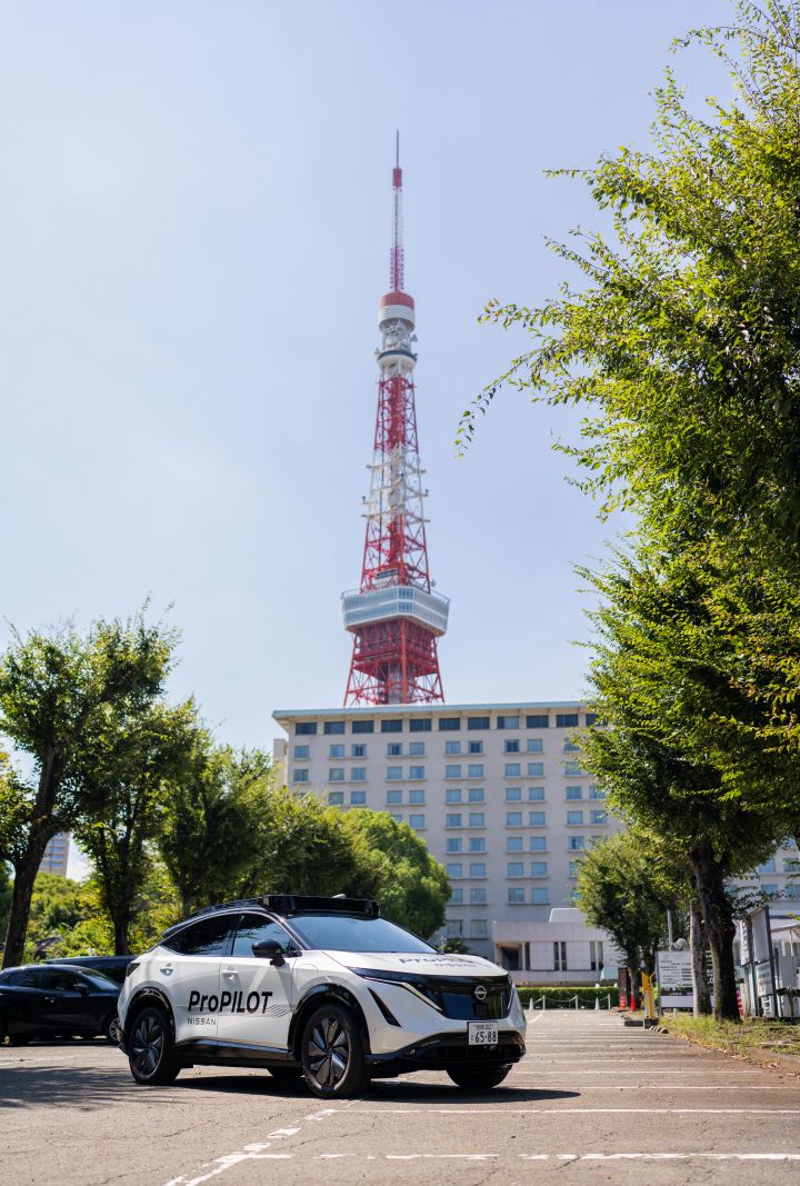 Ein weißer Nissan Ariya steht auf einem Parkplatz und ist nach links ausgerichtet. Im Hintergrund ragt der Tokyo Tower markant empor, unter ihm befindet sich ein großes Gebäude. Die Szene ist von grünen Bäumen umgeben, die die Natur mit der städtischen Landschaft verschmelzen lassen.