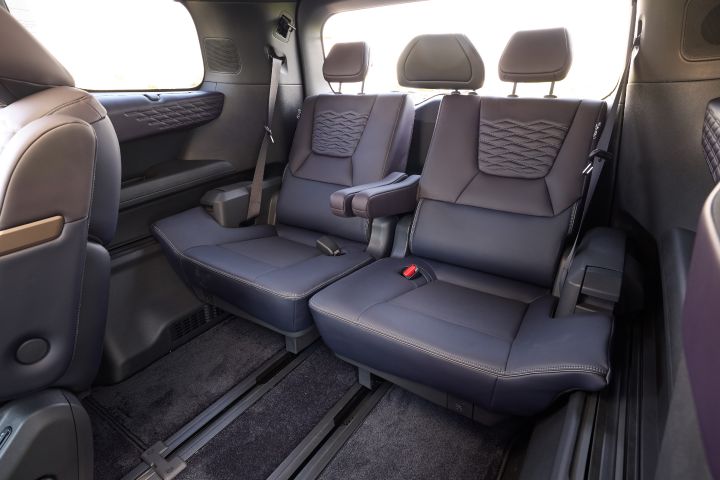 新型エルグランドの3列目シートを、車内前方から撮影した室内ビュー。左右に分割されたブラックのベンチシートはレザー調素材で、背もたれ部分にダイヤモンドステッチが施されている。各座席にはヘッドレストとシートベルトが備えられ、中央部分には折りたたまれたアームレストが見える。床面には前後スライド用レールが延び、足元にはカーペットが敷かれている。側面にはスピーカー付きの内装パネルと大きなウインドウがあり、外からの明るい日差しが差し込み、ゆとりある室内空間が表現されている。