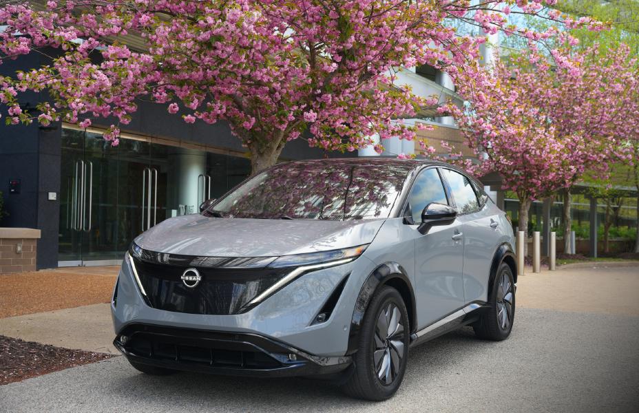 Front left 3/4 view of a gray 2024 Nissan Ariya parked under a tree with pink flowers.