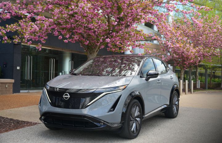 Front left 3/4 view of a gray 2024 Nissan Ariya parked under a tree with pink flowers.
