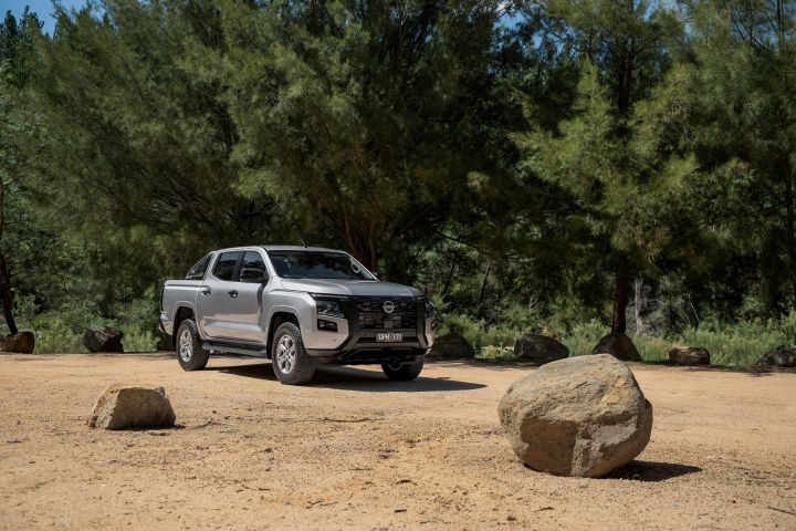 2026 Nissan Navara ST dual cab ute finished in Summit Silver shown from a distant front-left perspective in a sandy clearing with rocks and tall trees.