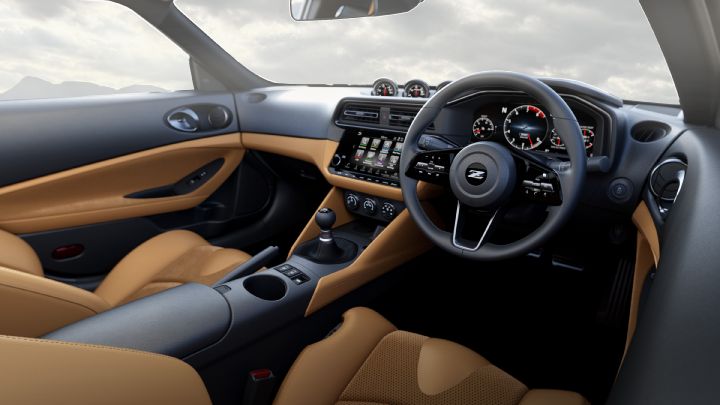 Interior view of a Fairlady Z from the driver’s side, focusing on the steering wheel and instrument cluster. The cabin features a two-tone design with camel brown seats and door panels contrasted by a black dashboard and center console. The steering wheel has a prominent “Z” logo at the center, with analog gauges and digital displays visible behind it. A touchscreen infotainment system and physical controls are positioned in the center, and the gear shift lever is located in the foreground. Through the windows, a soft gray sky and faint mountain silhouettes are visible, creating a calm and refined atmosphere.
