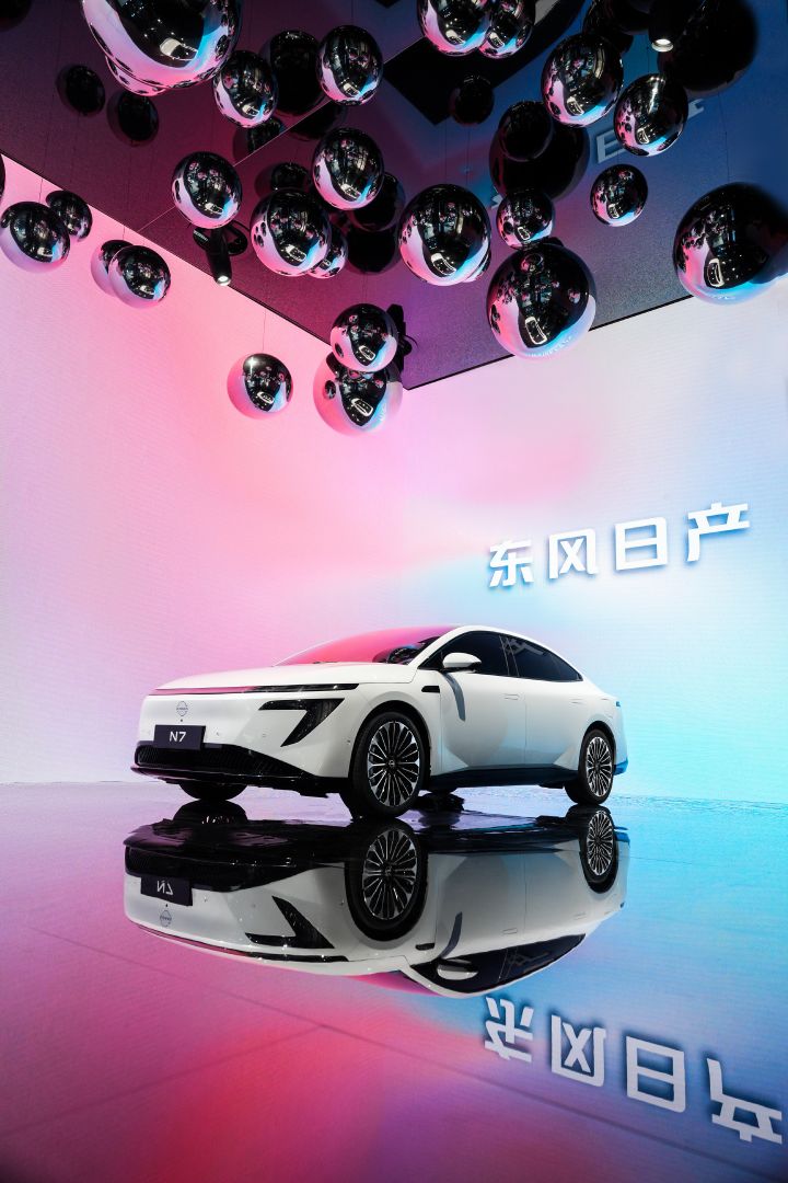 東風日産の白い新型セダン電気自動車「N7」のワイドショット。ピンクと青の背景、天井から銀色のボールが下がっている。