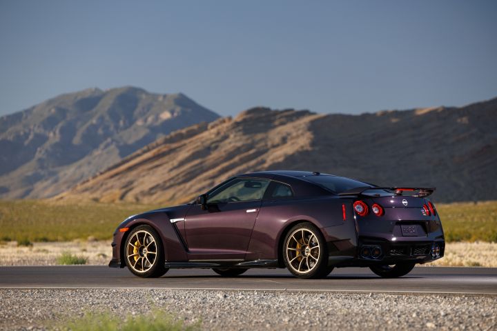 Left rear 3/4 angle of the Midnight Purple 2024 GT‑R T-spec Takumi parked on a track with grass and mountains behind.