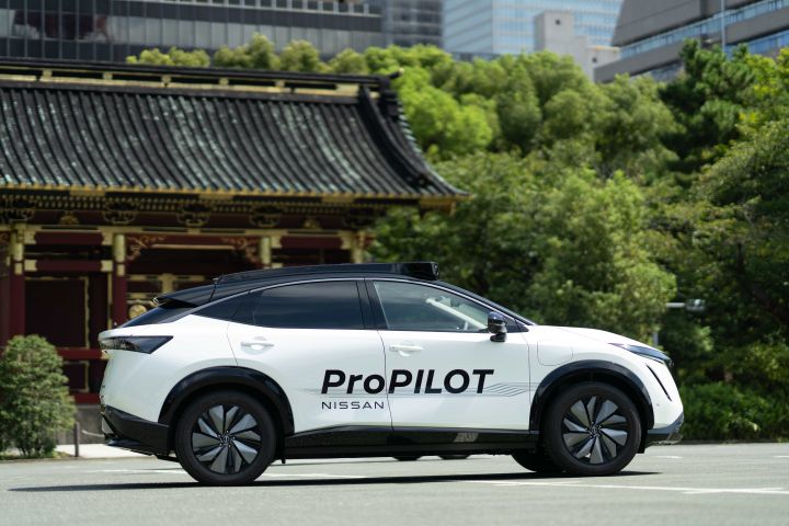 Ein weißer Nissan Ariya mit den schwarzen Logos „ProPILOT” und „NISSAN” steht seitlich vor einem großen Tempeltor, das von üppig grünen Bäumen umgeben ist, während sich in der Ferne moderne Hochhäuser erheben.