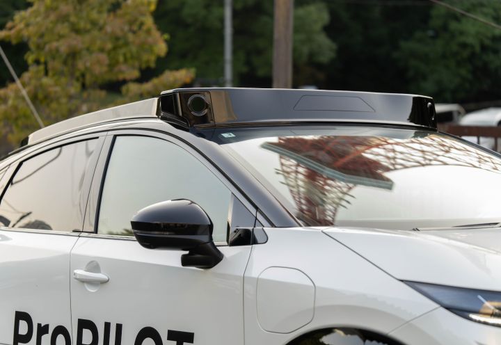 Detailaufnahme eines weißen Nissan Ariya, auf dessen Dach ein schwarzes Gerät zum autonomen Fahren montiert ist. Im Hintergrund sind grüne Bäume und ein Strommast zu sehen.