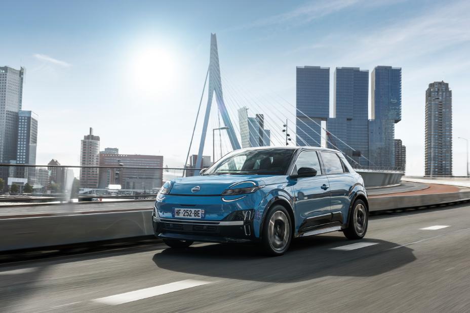 3/4-Front-Fahraufnahme eines blauen Nissan Micra auf eienr Brücke vor mehreren Hochhäusern im Zentrum von Rotterdam
