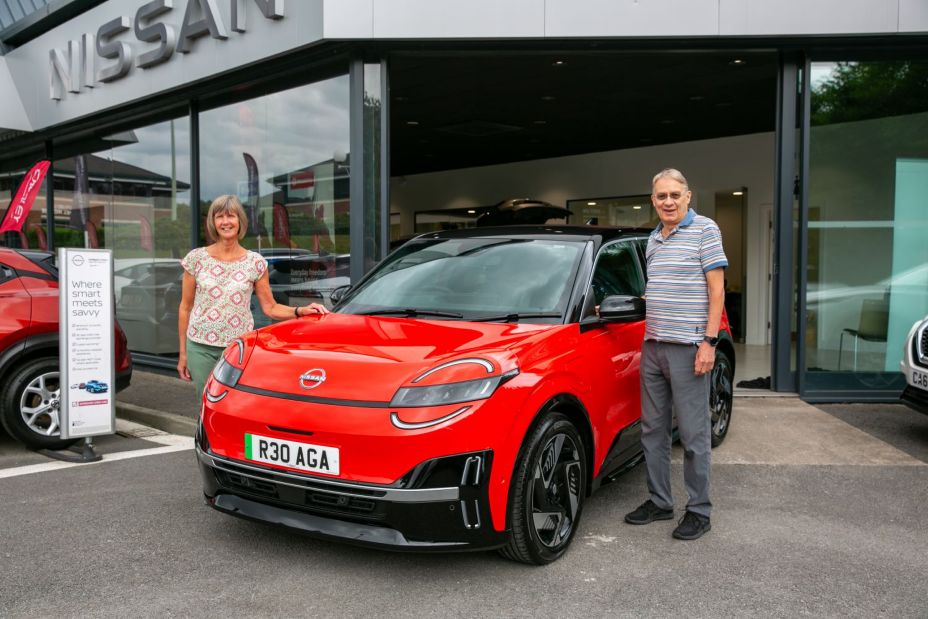 Photo of a retired couple standing next to their new bright red Nissan MICRA – the 100,000th Nissan EV to be sold in the UK