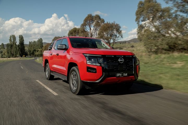 Front three‑quarter close-up of a 2026 Nissan Navara ST‑X Outback Red dual cab, driving on a sealed rural road edged by tall trees and roadside grass.