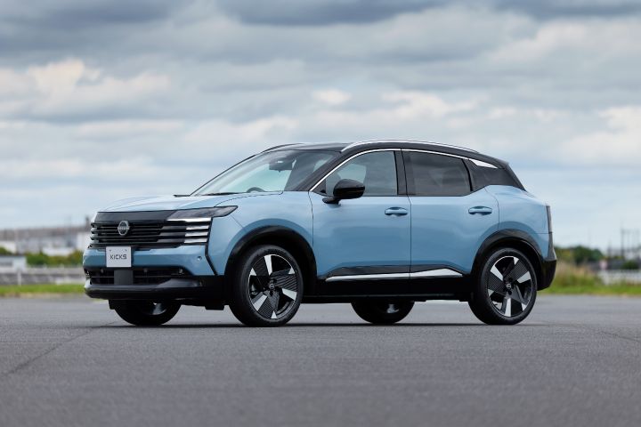 青みがかったキックスを低い位置から捉えた斜め前方ビュー。車両は右向きに配置され、背景には広いアスファルト路面と曇り空が広がる。