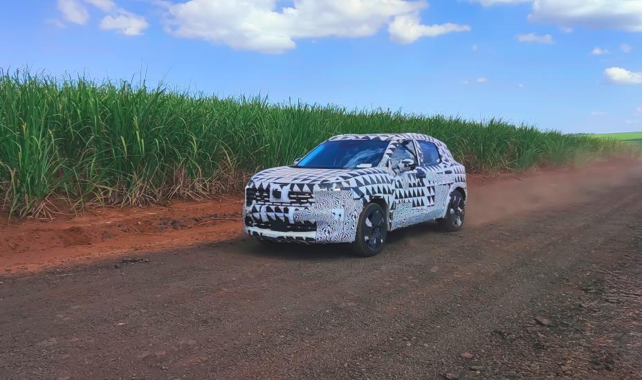 Unidade do SUV camuflada trafega por estrada de terra cercada por vegetação alta, levantando poeira sob céu azul com nuvens