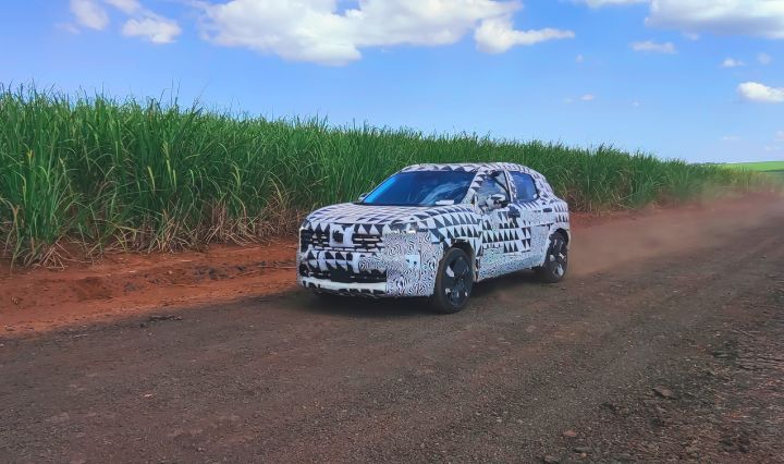 Unidade do SUV camuflada trafega por estrada de terra cercada por vegetação alta, levantando poeira sob céu azul com nuvens