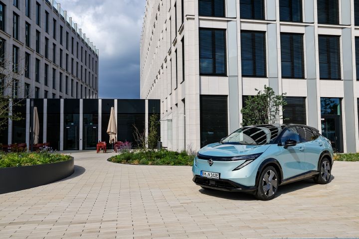 Auf einem hellen, sonnenbeschienenen Platz steht schräg ein hellblauer Nissan Ariya mit einem modernen Bürogebäude im Hintergrund
