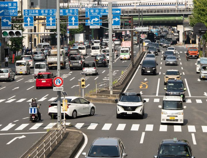 Ein weißer Nissan Ariya fährt auf einer großen, vielbefahrenen Kreuzung vorwärts. Auf der Straße befinden sich mehrere Fahrzeuge, und im Hintergrund sind blaue Verkehrsschilder, Gebäude und eine Überführung zu sehen, die eine städtische Szenerie darstellen.