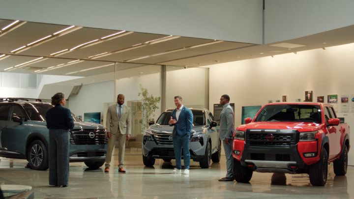 A  group of people in business clothes, standing around three Nissan vehicles in a showroom.