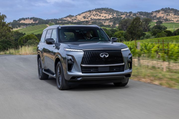ブドウ畑と木々を背に田舎道を走るダークグレーの INFINITI QX80 SUV の正面 3/4 ビュー