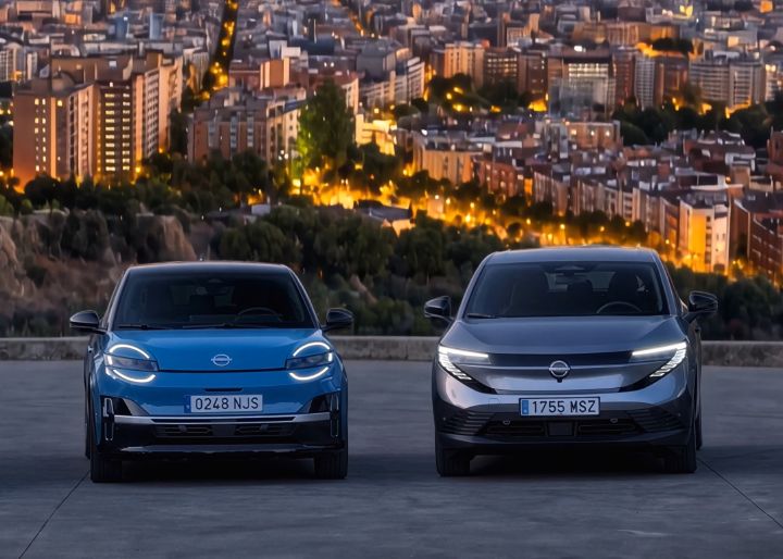 Nissan Micra elétrico azul ao lado de novo Nissan LEAF cinzento, vistos de frente, com cidade de Barcelona em background