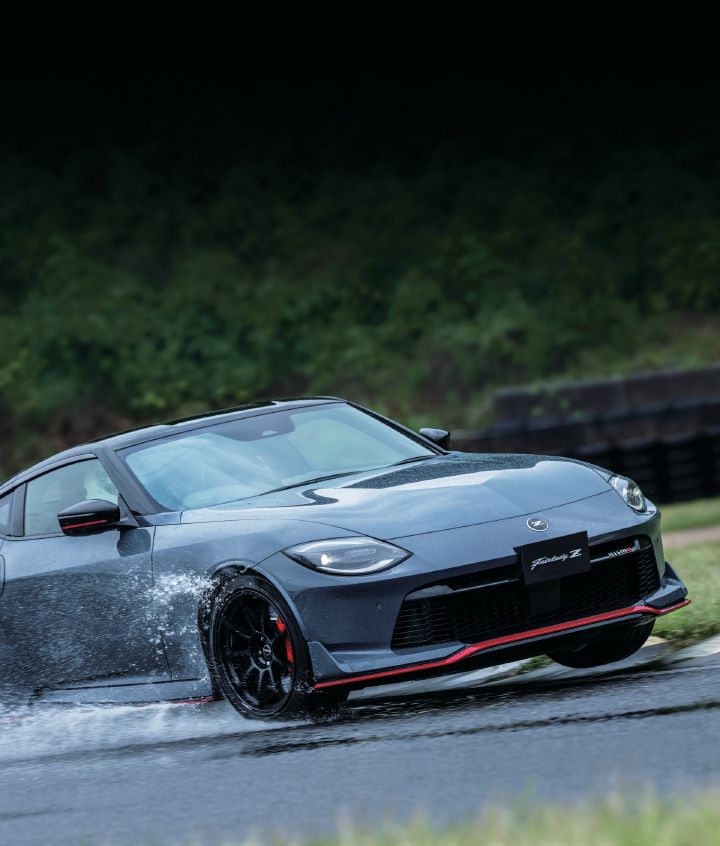 Gray Fairlady Z NISMO driving on a rain-soaked race circuit, photographed from a front-right three-quarter angle with the vehicle facing toward the right side of the image. The car features a NISMO-specific front bumper, large grille, and red accent trim along the front splitter and side sills. Black alloy wheels and red brake calipers emphasize its performance-focused character. Water sprays dramatically from the front wheel area as the vehicle passes through standing water on the wet track surface. Tire barriers and green vegetation fill the background, creating a dynamic rainy-weather scene that highlights the handling capability and aggressive styling of the Fairlady Z NISMO.