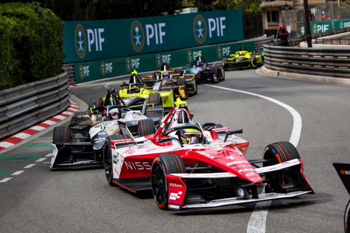 Oliver Rowland auf der Rennstrecke im Formel E Rennwagen mit der Startnummer 1, hinter ihm mehrere andere Autos und im Hintergrund grüne Werbetafeln.