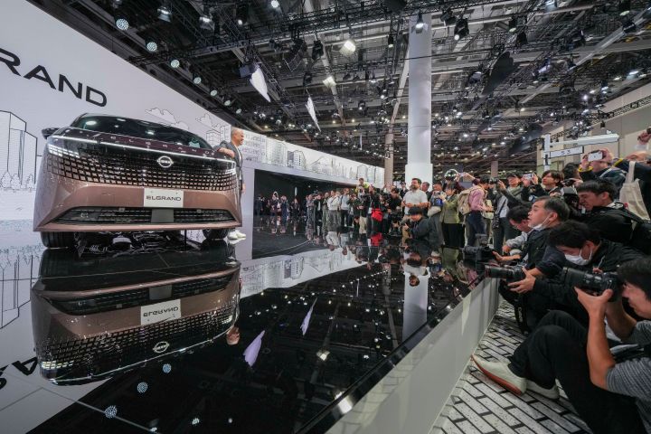 On the stage, the all-new Elgrand in Fuji Dawn and Shigoku two-tone colors is positioned diagonally, facing slightly to the left.  In the background, a white-themed screen features Elgrand logo. On the right side of the foreground, members of the press are gathered, taking photos with their cameras.